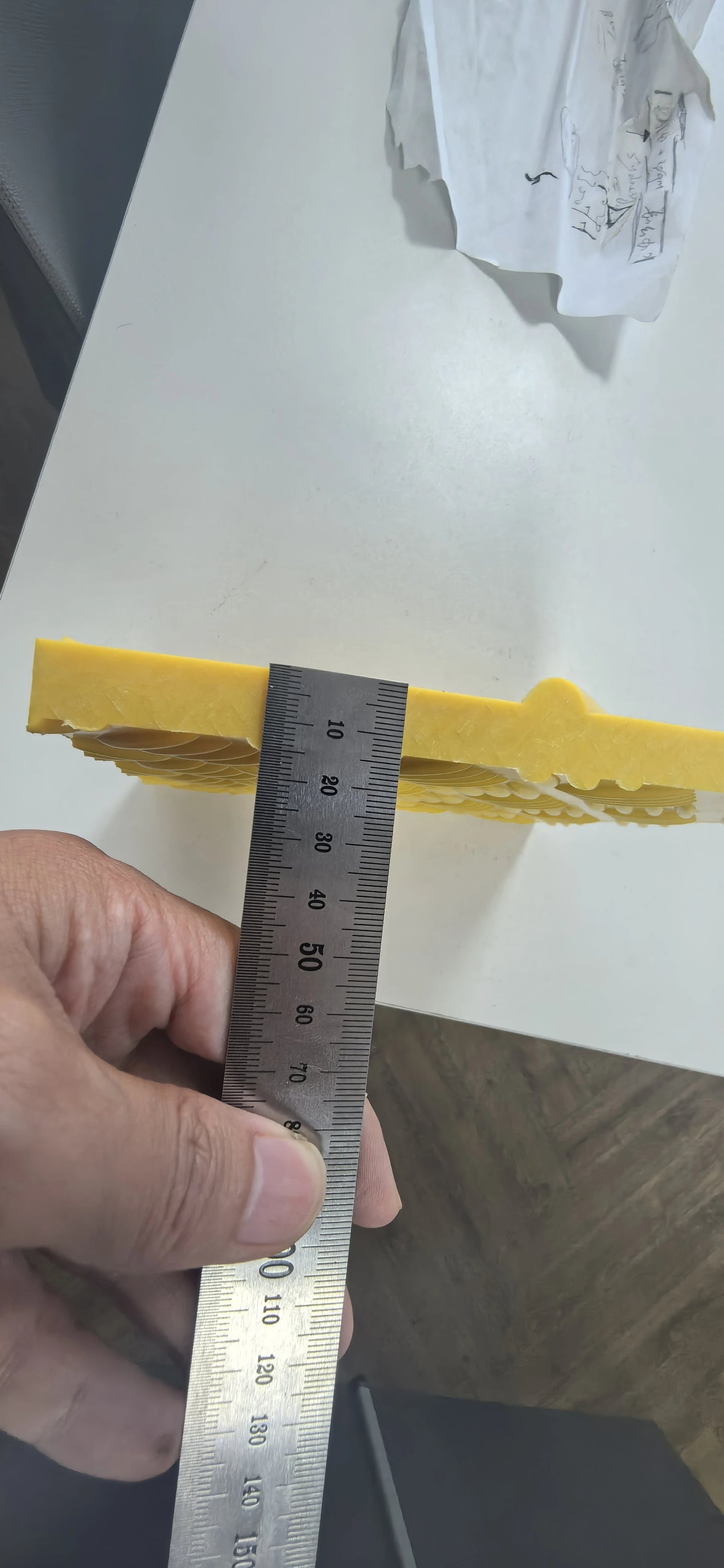 Hand holding a metal ruler against a yellow object on a white surface.
