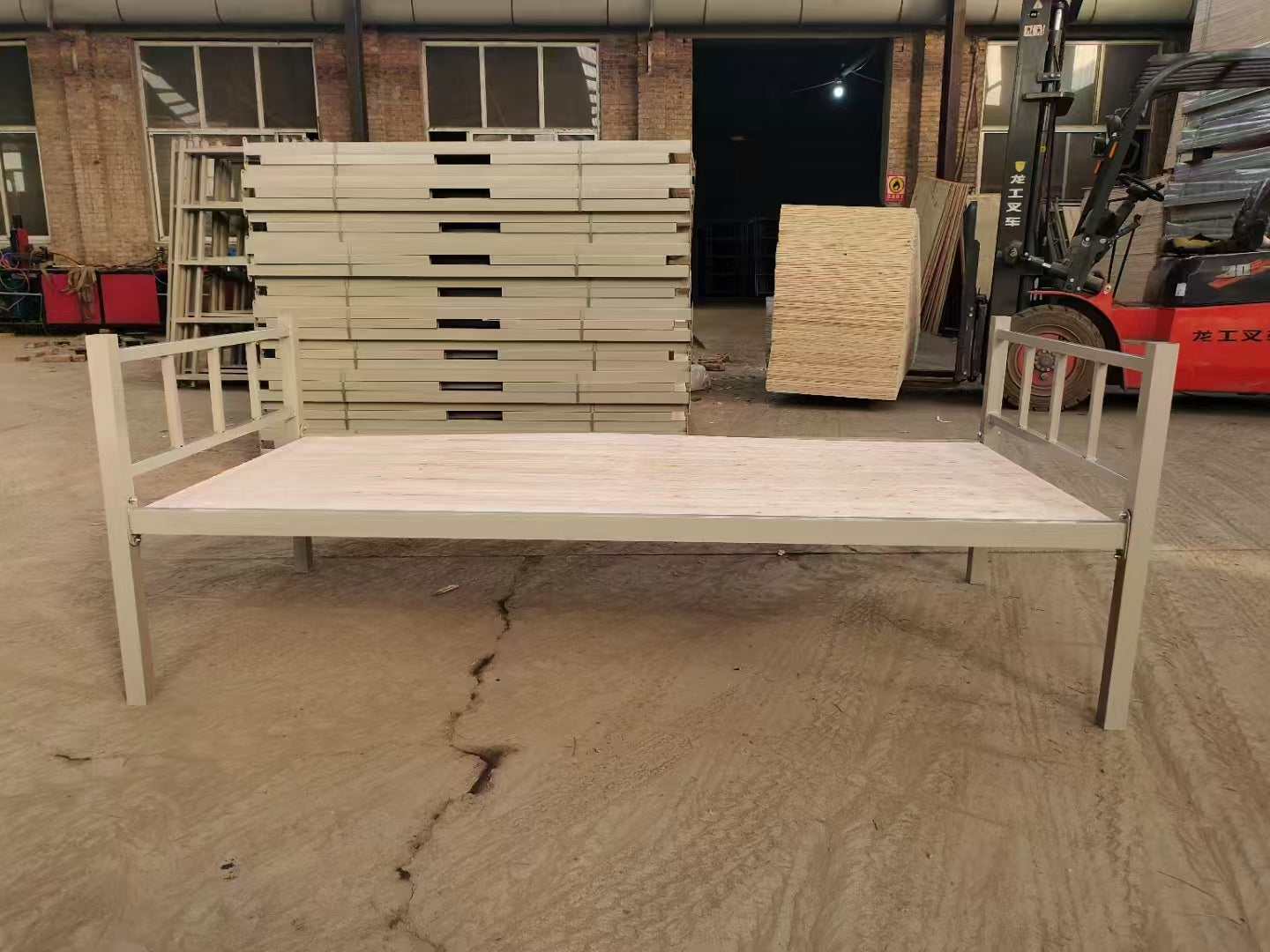 Metal bed frame on a concrete floor with stacks of wooden panels in the background.