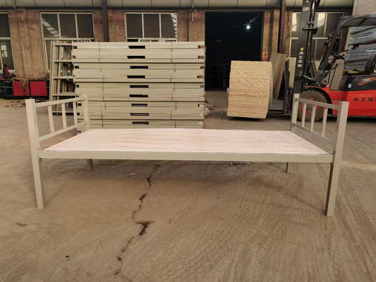 Metal bed frame on a concrete floor with stacks of wooden panels in the background.